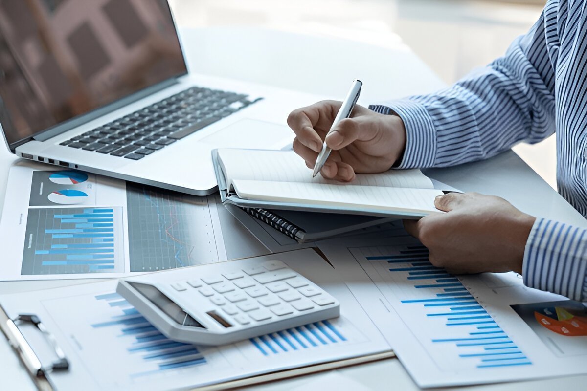 Person taking notes beside a laptop, calculator and printed revenue charts